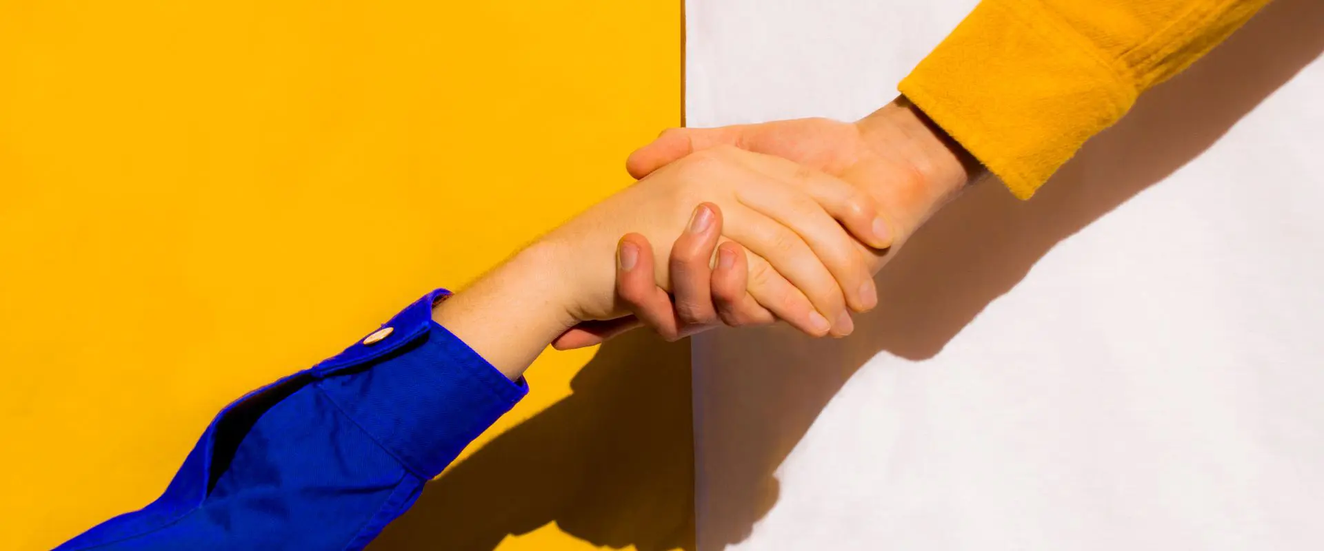 In the image there are two hands shaking. Both people are wearing shirts, the one on the left is blue and the one on the right is yellow. The background is divided in half yellow and white.
